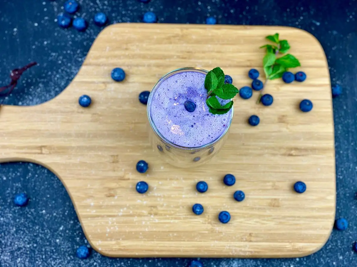 Vaso de smoothie de arándanos lila con menta sobre una tabla de madera rodeada de arándanos sueltos