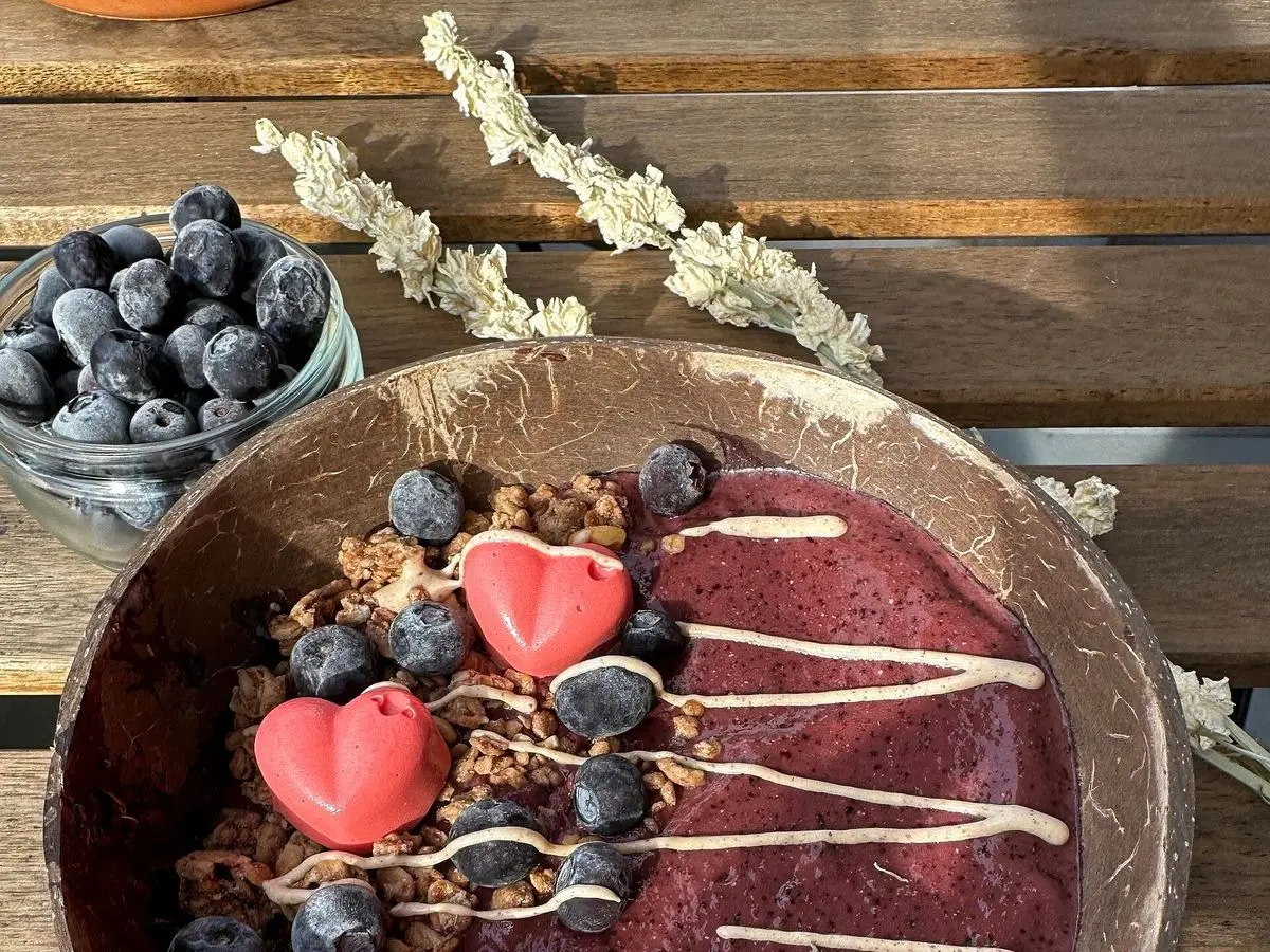 Cuenco de smoothie bowl morado con arándanos, corazones de fruta, granola y hilos de salsa blanca