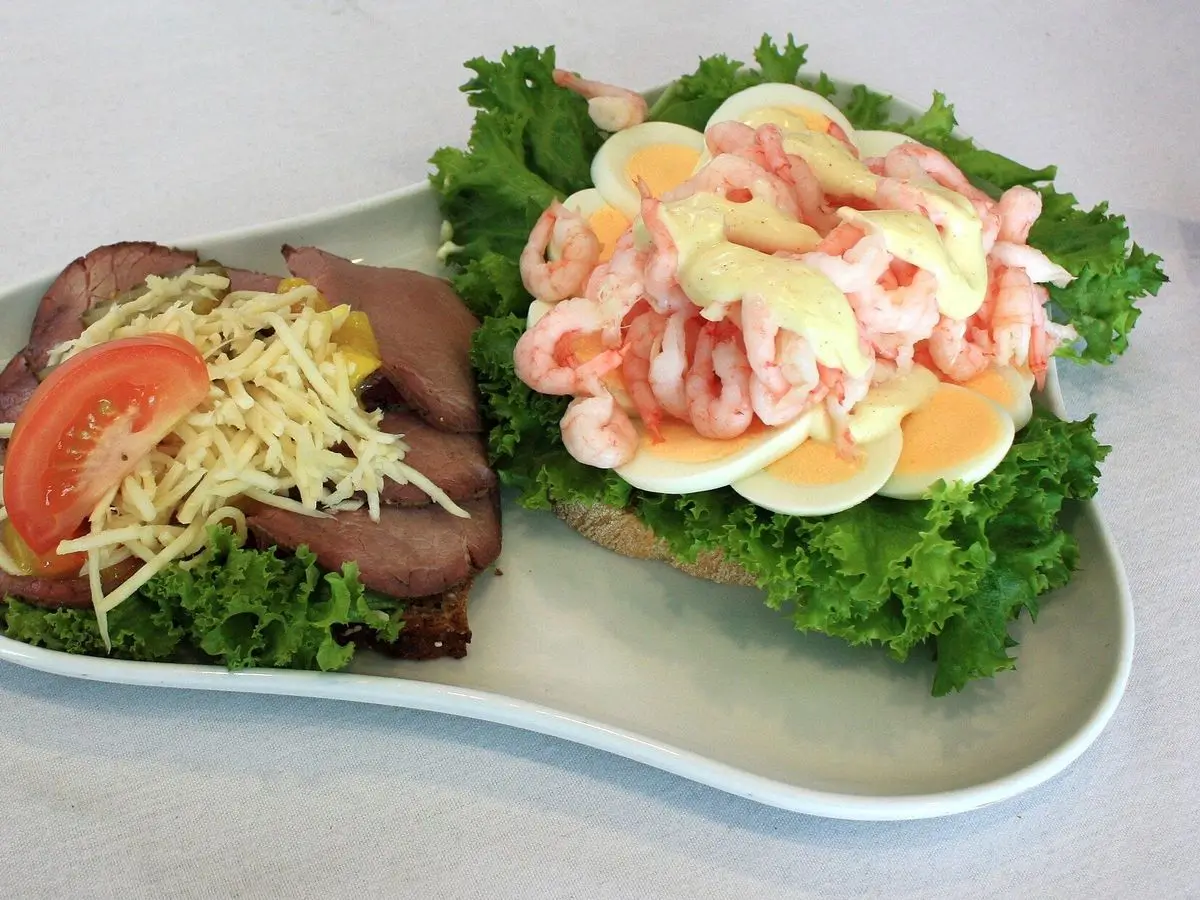 Tostada abierta de pan con gambas peladas, huevo cocido y lechuga junto a otro bocado con rosbif