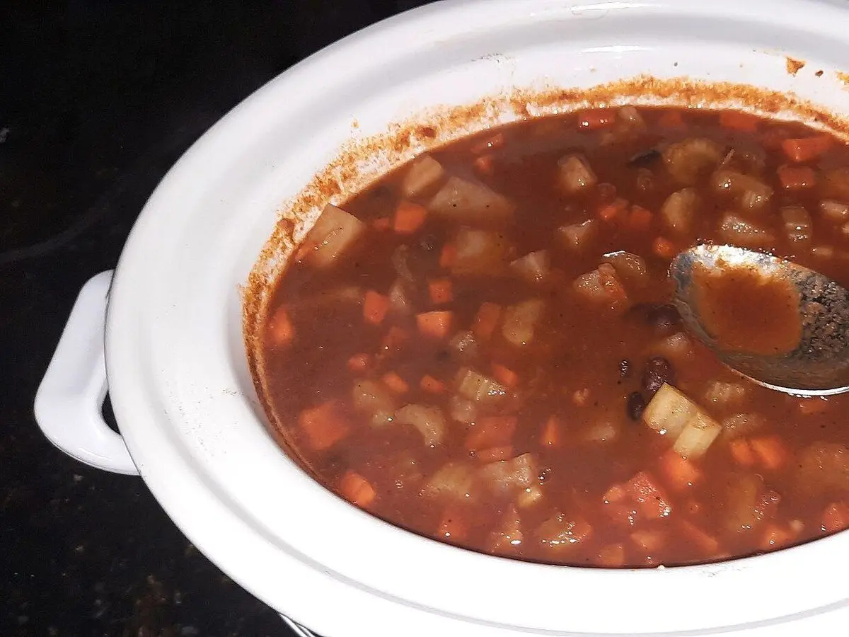 Olla blanca con guiso vegetal de patata, legumbres y zanahoria en caldo rojo y una cuchara