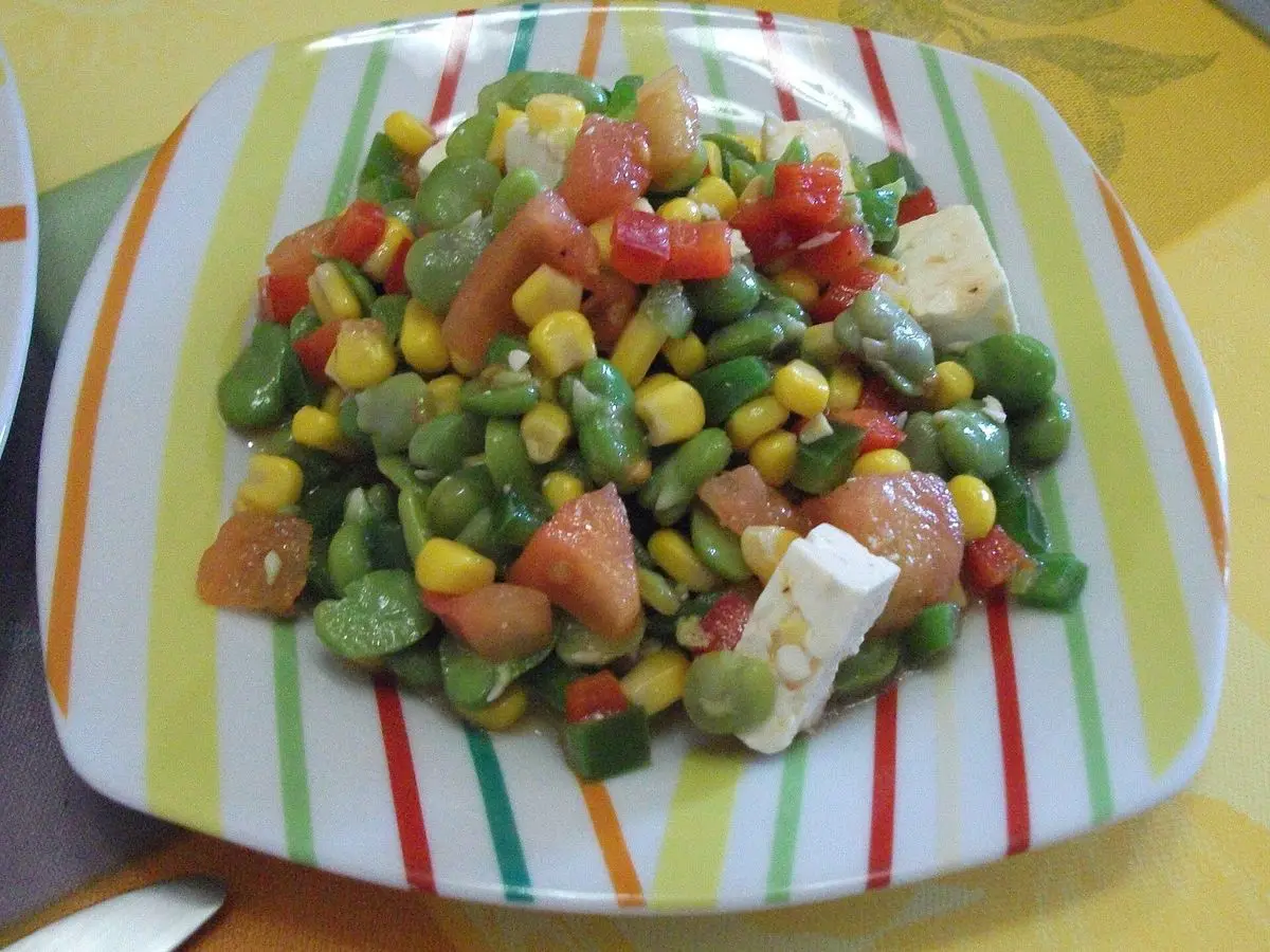 Plato con ensalada de habas, maíz, tomate y queso fresco en cubos sobre mantel de rayas