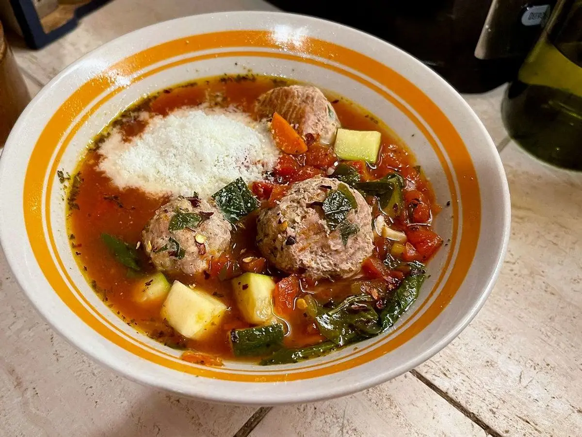 Plato de sopa de albóndigas con tomate, verduras, hierbas y queso rallado