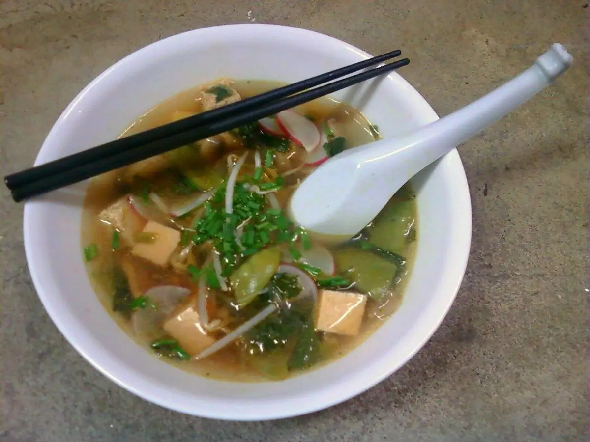 Cuenco blanco de sopa asiática con dados de tofu, verduras, hierbas y fideos, con cuchara y palillos