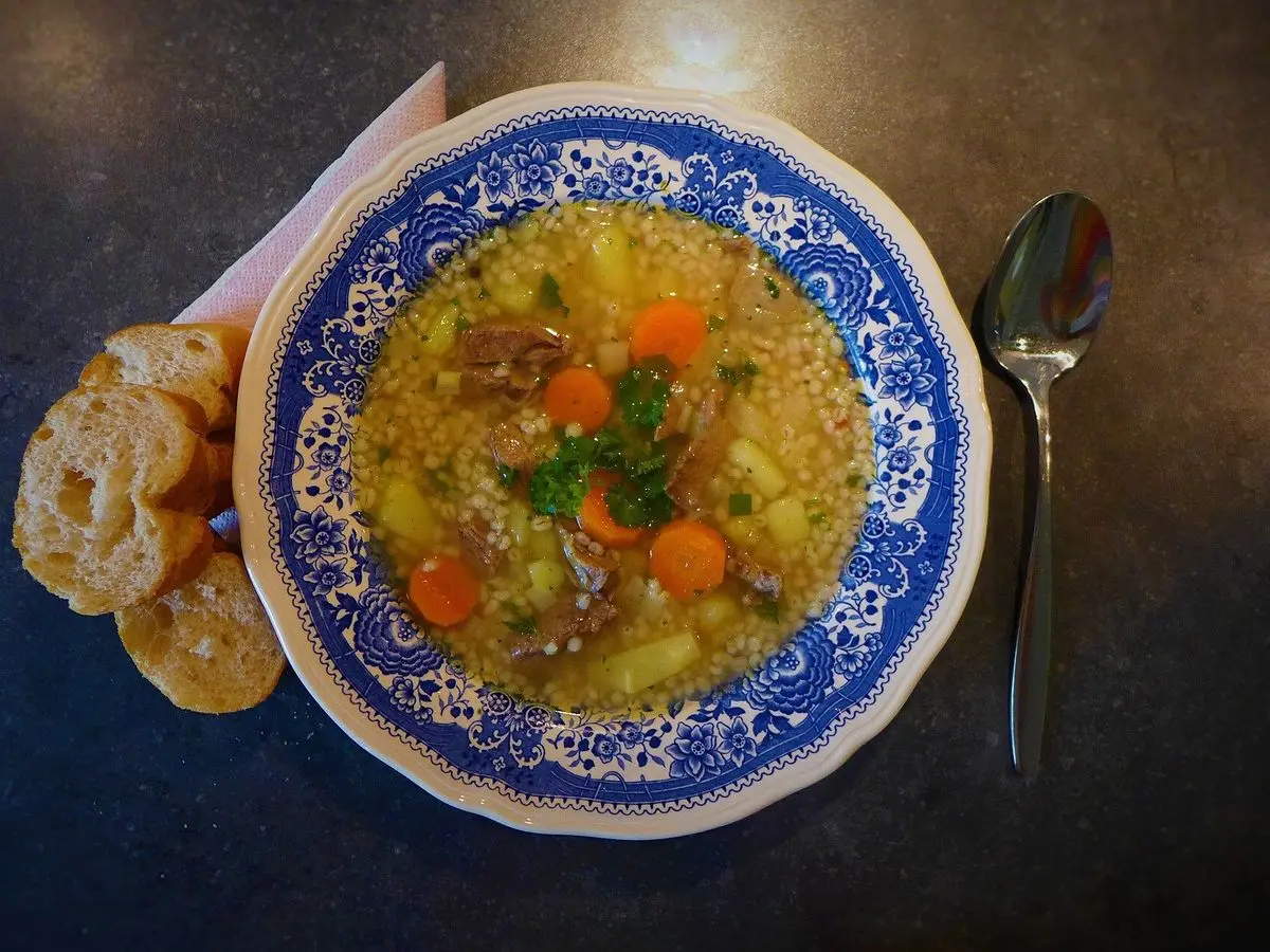 Plato de sopa de cebada perlada con carne, zanahoria y verduras junto a trozos de pan y una cuchara