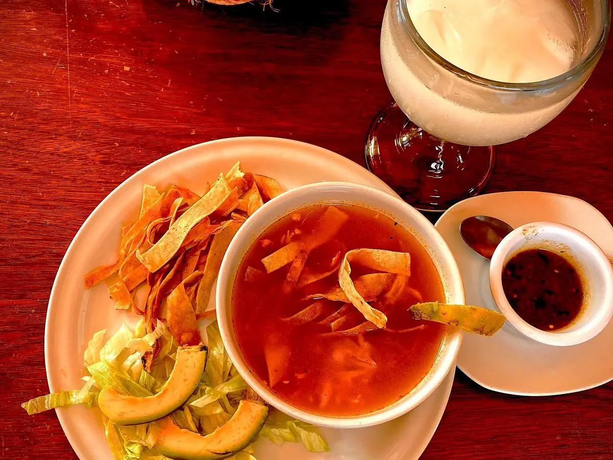 Cuenco blanco de sopa anaranjada con tiras de tortilla frita, aguacate y lechuga en un plato, sobre mesa de madera roja
