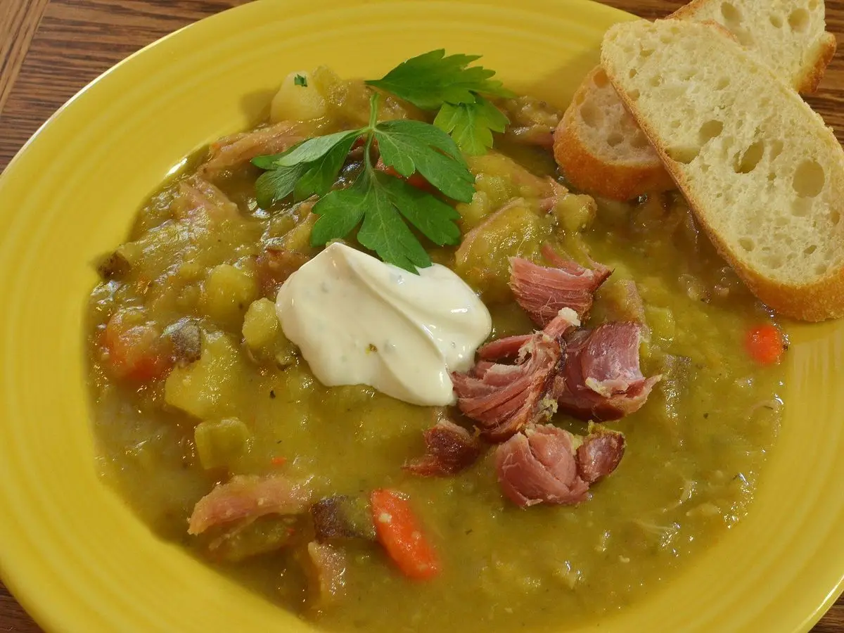 Plato amarillo de sopa de guisantes partidos con trozos de jamón, patata y zanahoria con pan