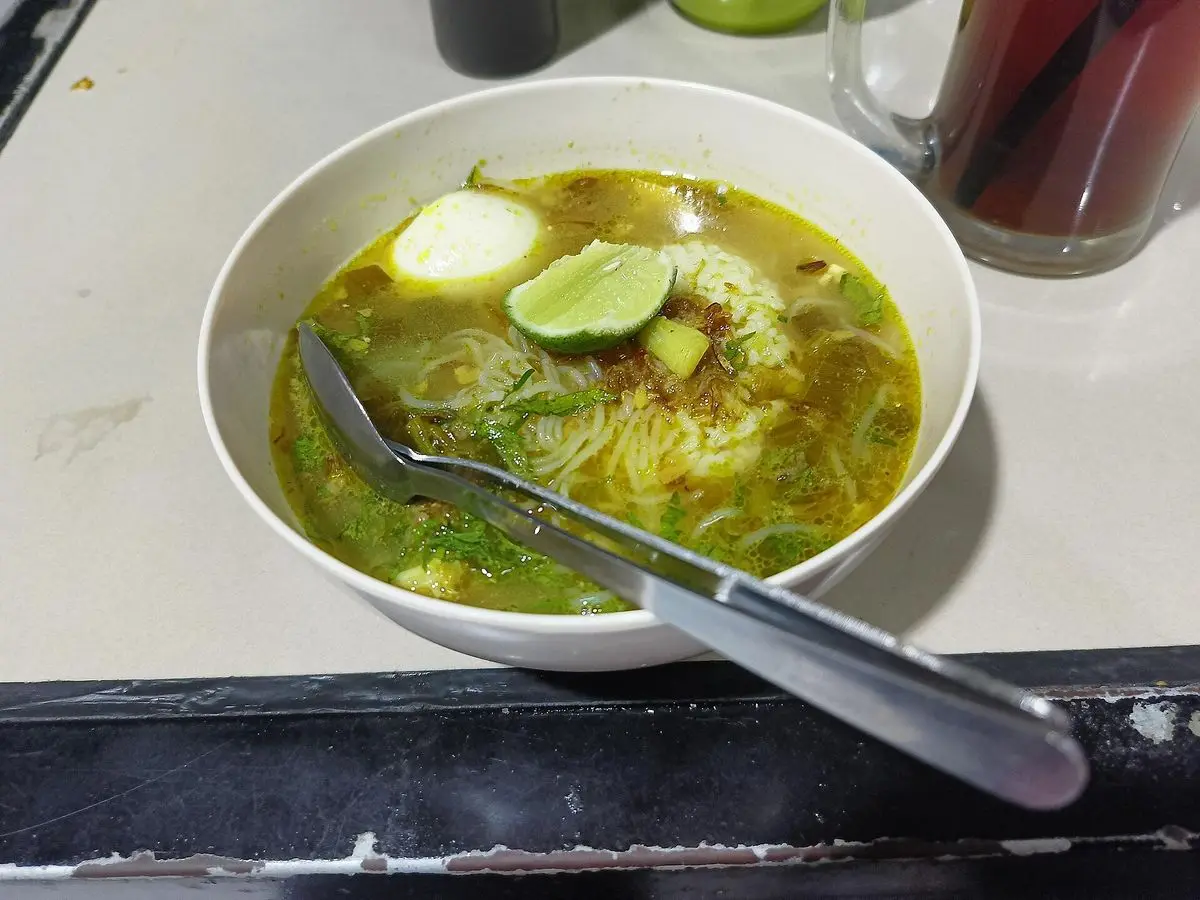 Cuenco de sopa amarilla de pollo con huevo cocido, lima y cuchara metálica