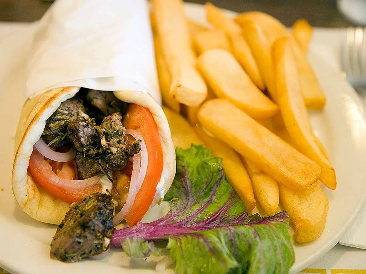 Pan de pita enrollado con carne, tomate y cebolla morada junto a patatas fritas y hojas verdes