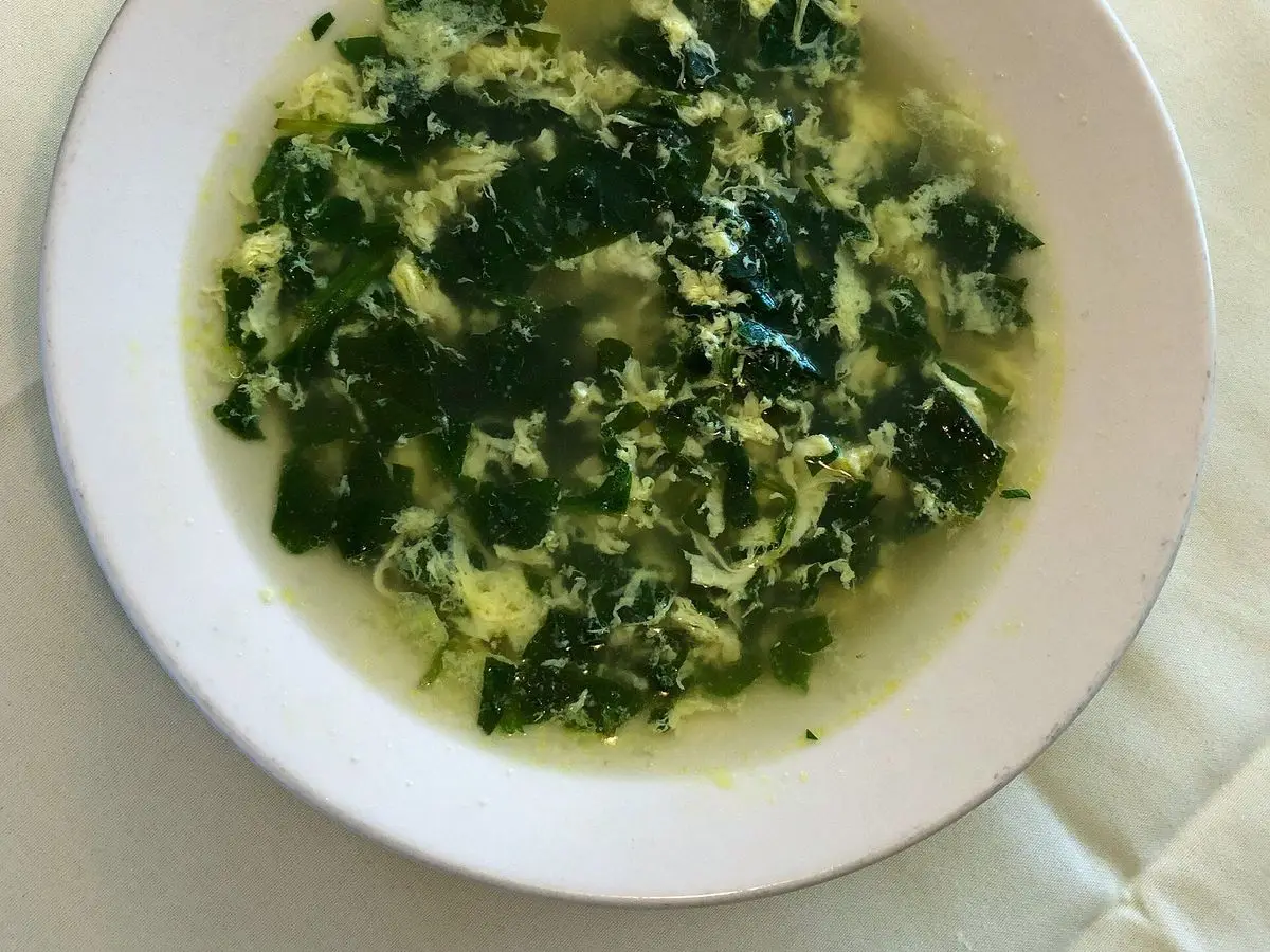 Plato hondo blanco de sopa stracciatella con espinacas y huevo en hilos sobre un mantel claro
