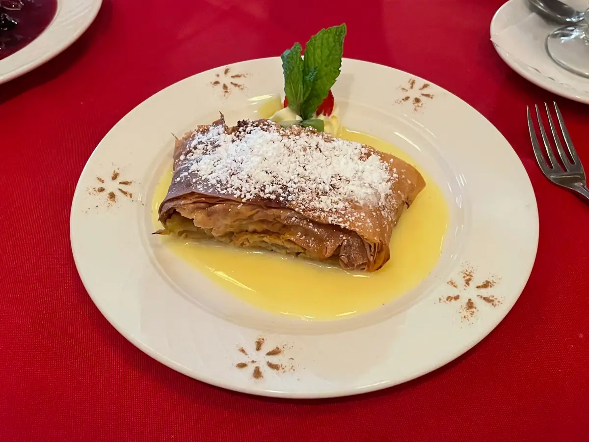 Porción de strudel de manzana espolvoreada de azúcar glas sobre salsa de vainilla en un plato con hoja de menta