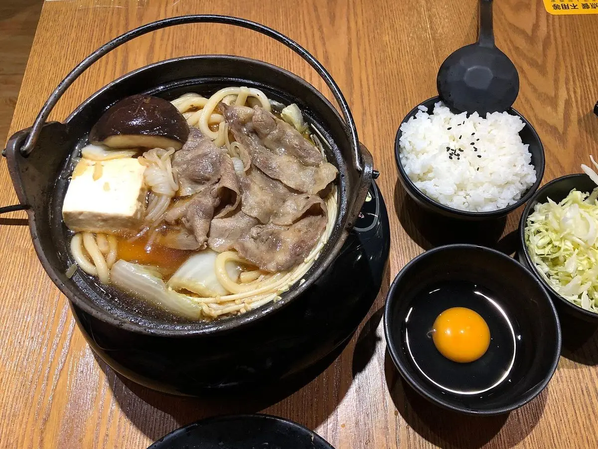 Olla de hierro con sukiyaki de carne y tofu, junto a arroz blanco y un huevo crudo