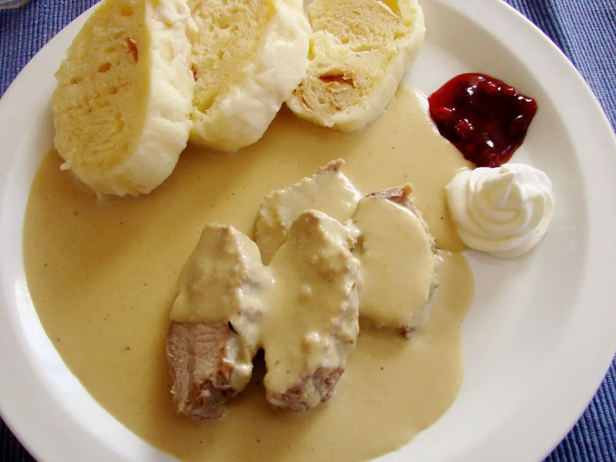 Lonchas de carne en salsa cremosa con rodajas de knedlík, arándanos rojos y nata montada