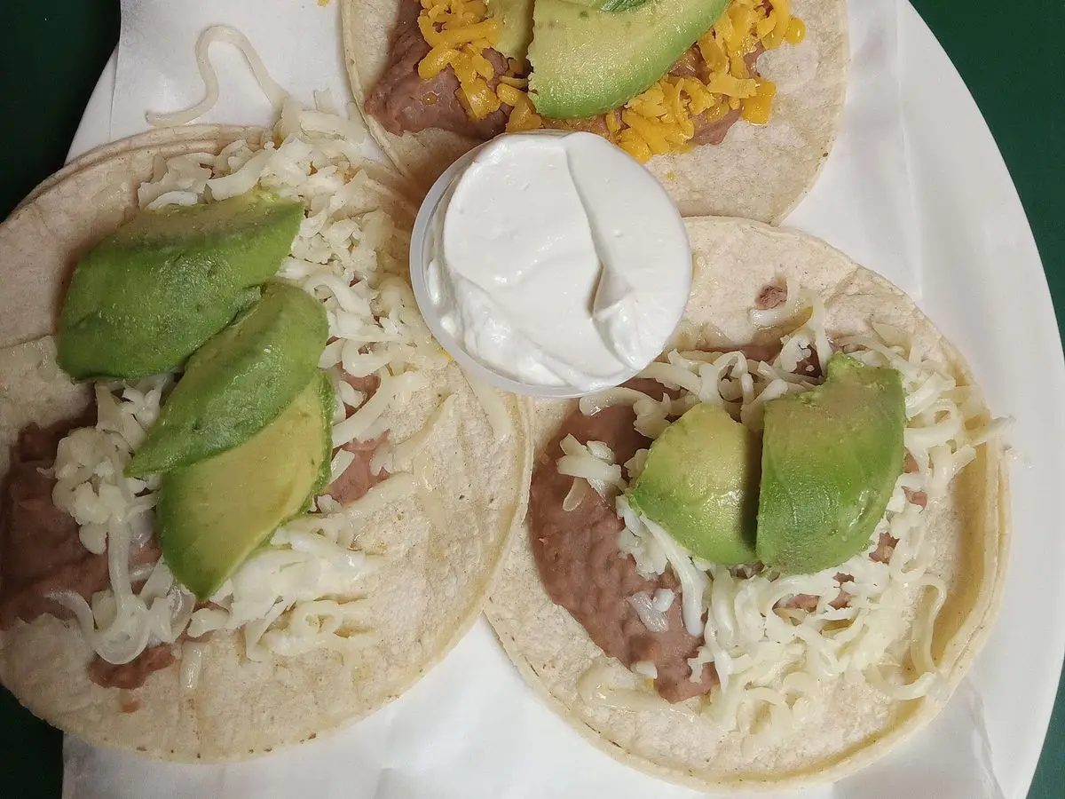 Tres tacos de tortilla de maíz con frijoles, aguacate en láminas, queso rallado y crema agria