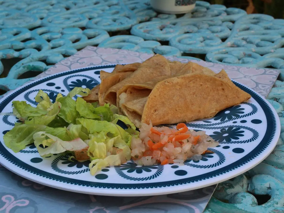 Tacos dorados de papa con lechuga y salsa roja en un plato decorado sobre una mesa de forja turquesa
