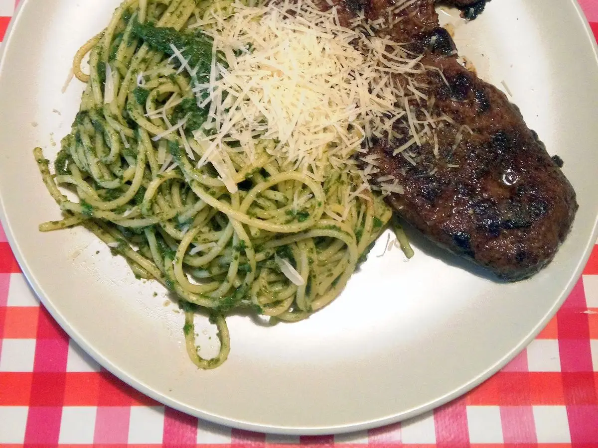 Plato con espaguetis verdes al pesto con queso rallado y un filete de ternera a la plancha sobre mantel de cuadros