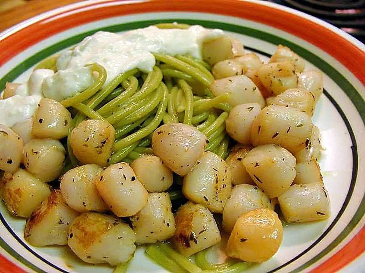 Plato de pasta verde con vieiras pequeñas doradas y una cucharada de crema blanca