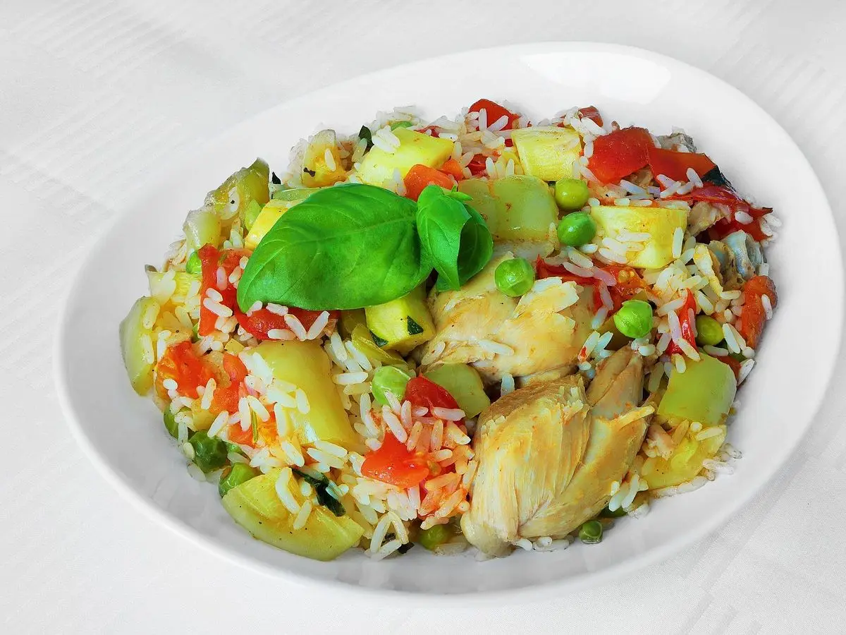 Plato de arroz con pollo, pimientos, tomate, guisantes y calabacín decorado con una hoja de albahaca