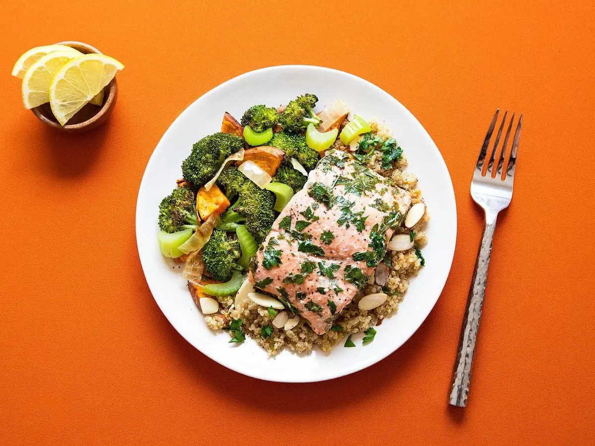 Plato de salmón al horno con brócoli y quinoa sobre fondo naranja, con limones y un tenedor
