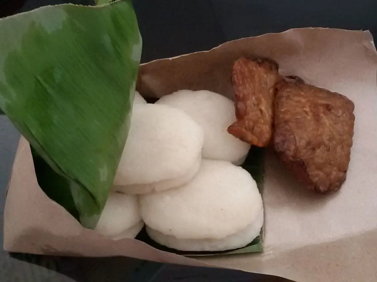 Tempeh frito marrón junto a bolitas blancas de arroz sobre hoja de plátano y papel