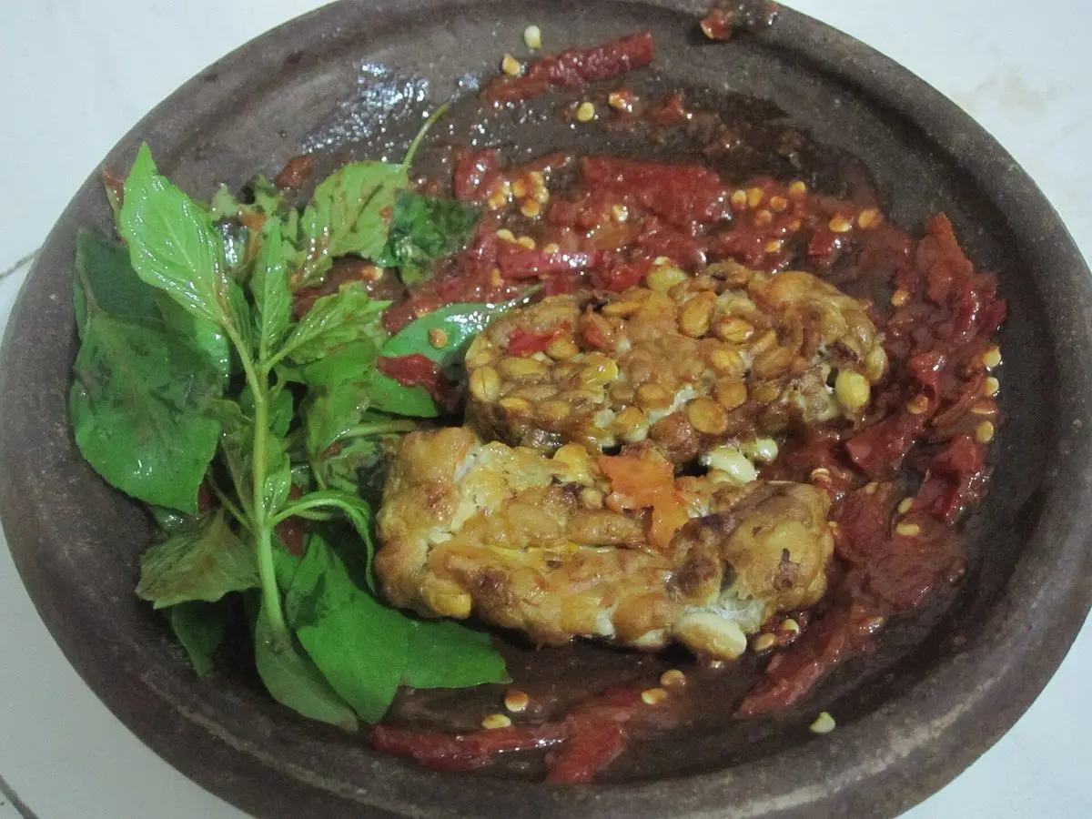 Tempeh frito aplastado sobre sambal rojo picante en un mortero de piedra, con hojas de albahaca fresca