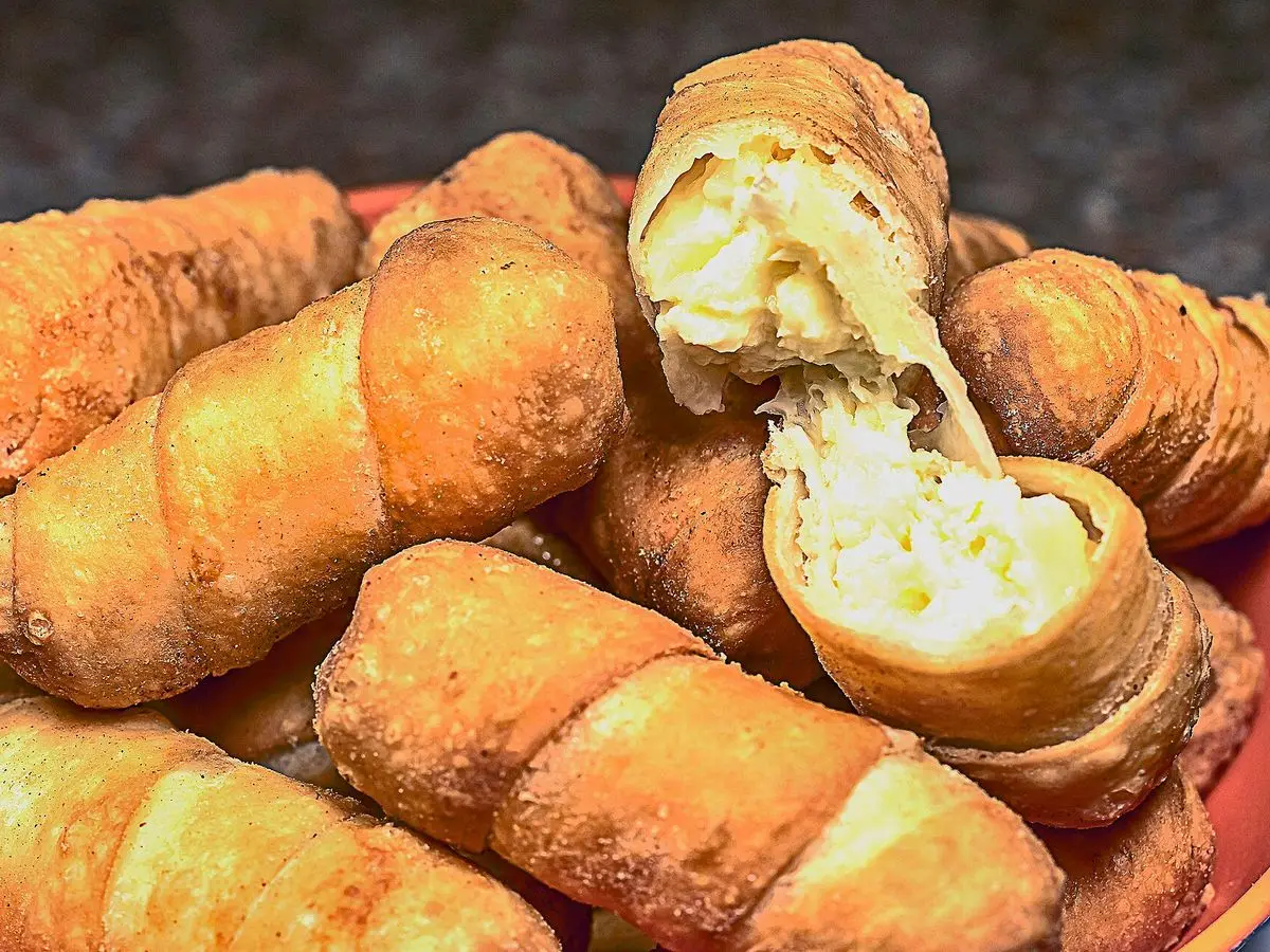 Tequeños dorados apilados, uno abierto que deja ver el queso fundido del relleno