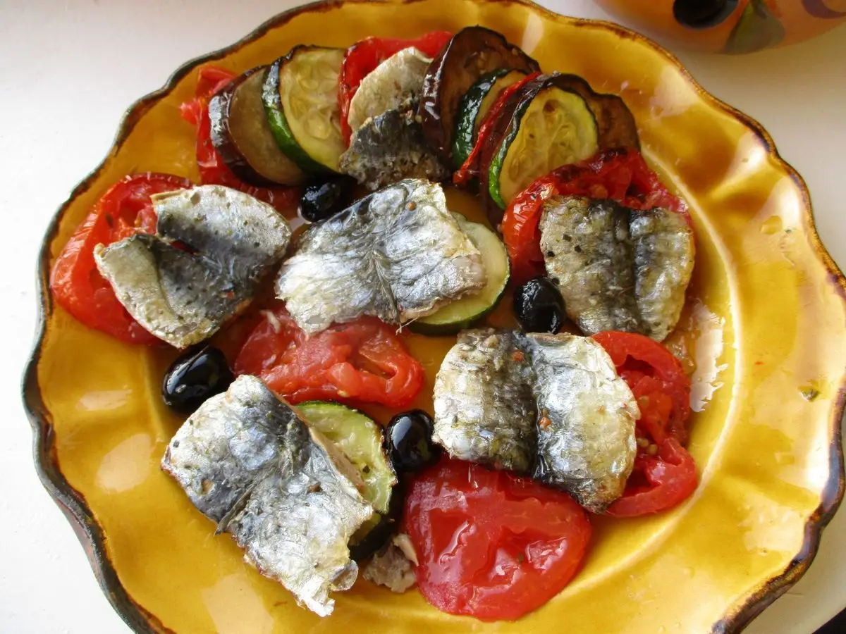 Fuente amarilla con filetes de sardina sobre verduras al horno: tomate, calabacín, berenjena y aceitunas