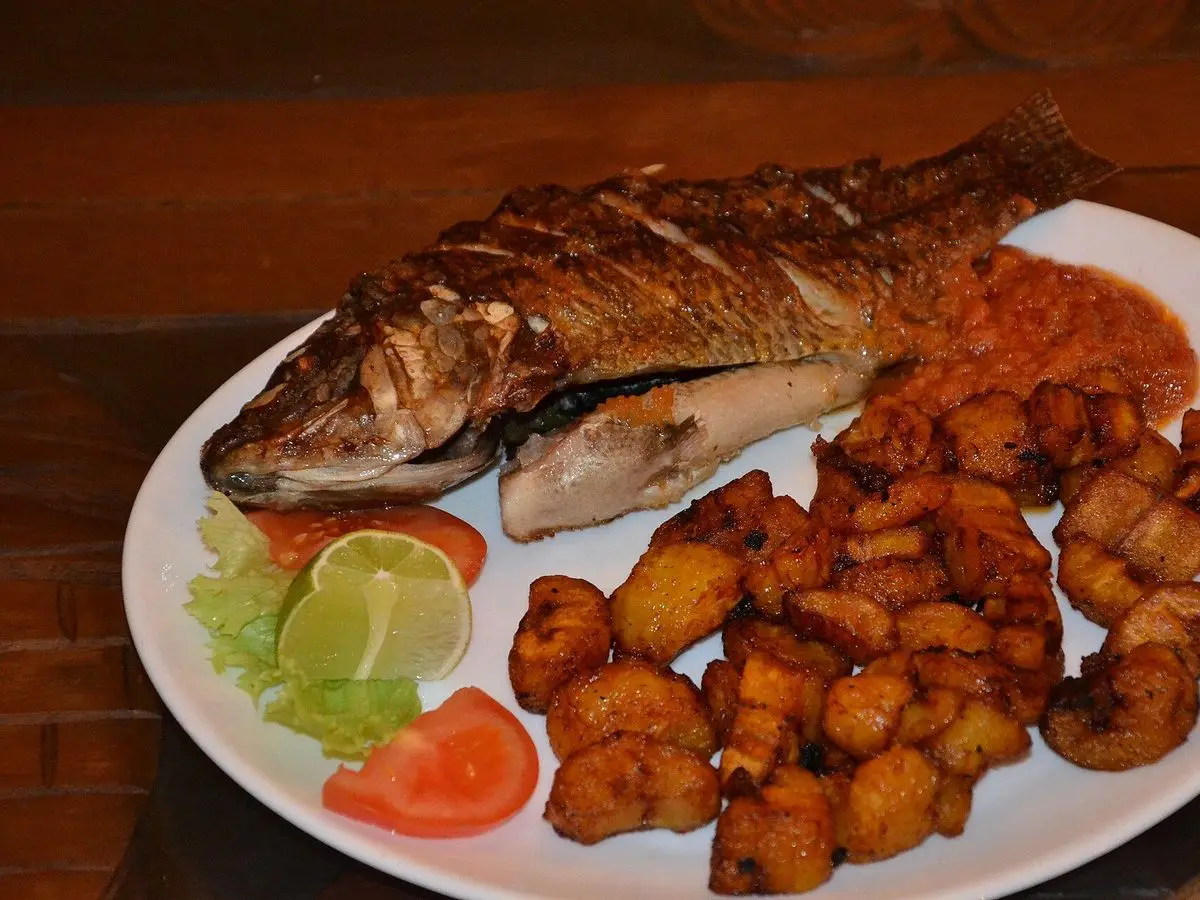 Pescado entero asado con plátano macho frito en trozos, rodaja de tomate y lima en un plato blanco