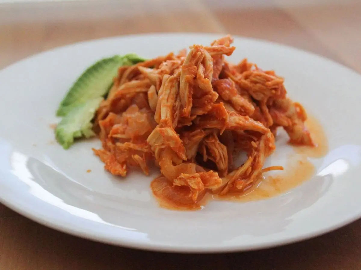 Pollo deshebrado en salsa roja de tomate y chipotle con rodaja de aguacate en un plato blanco