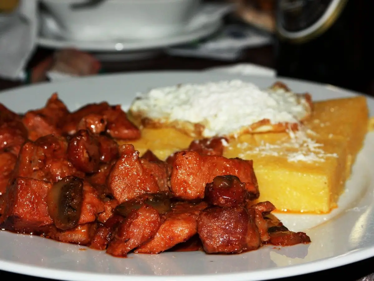 Trozos de cerdo en salsa roja con polenta y queso rallado en un plato blanco