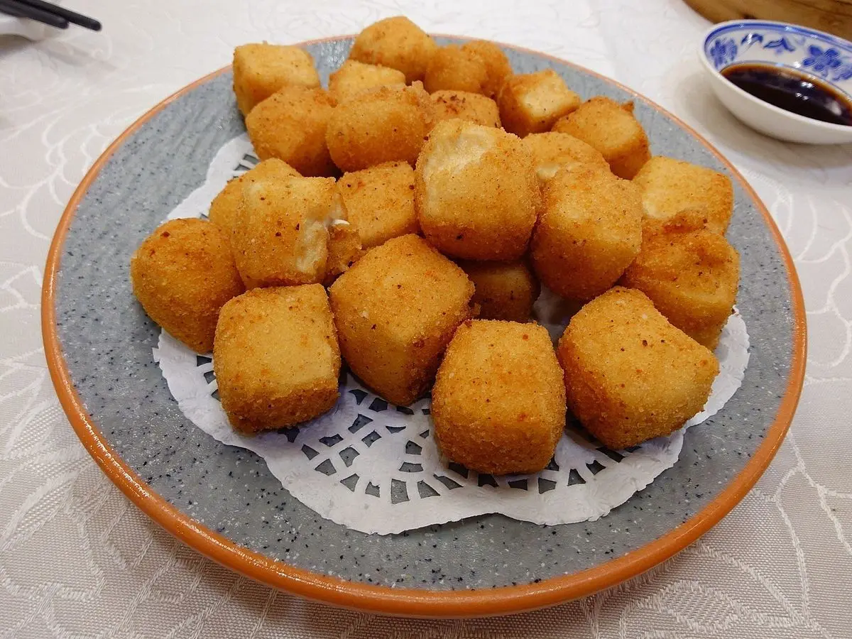 Dados de tofu rebozados y dorados servidos en un plato sobre papel calado, con salsa de soja al fondo