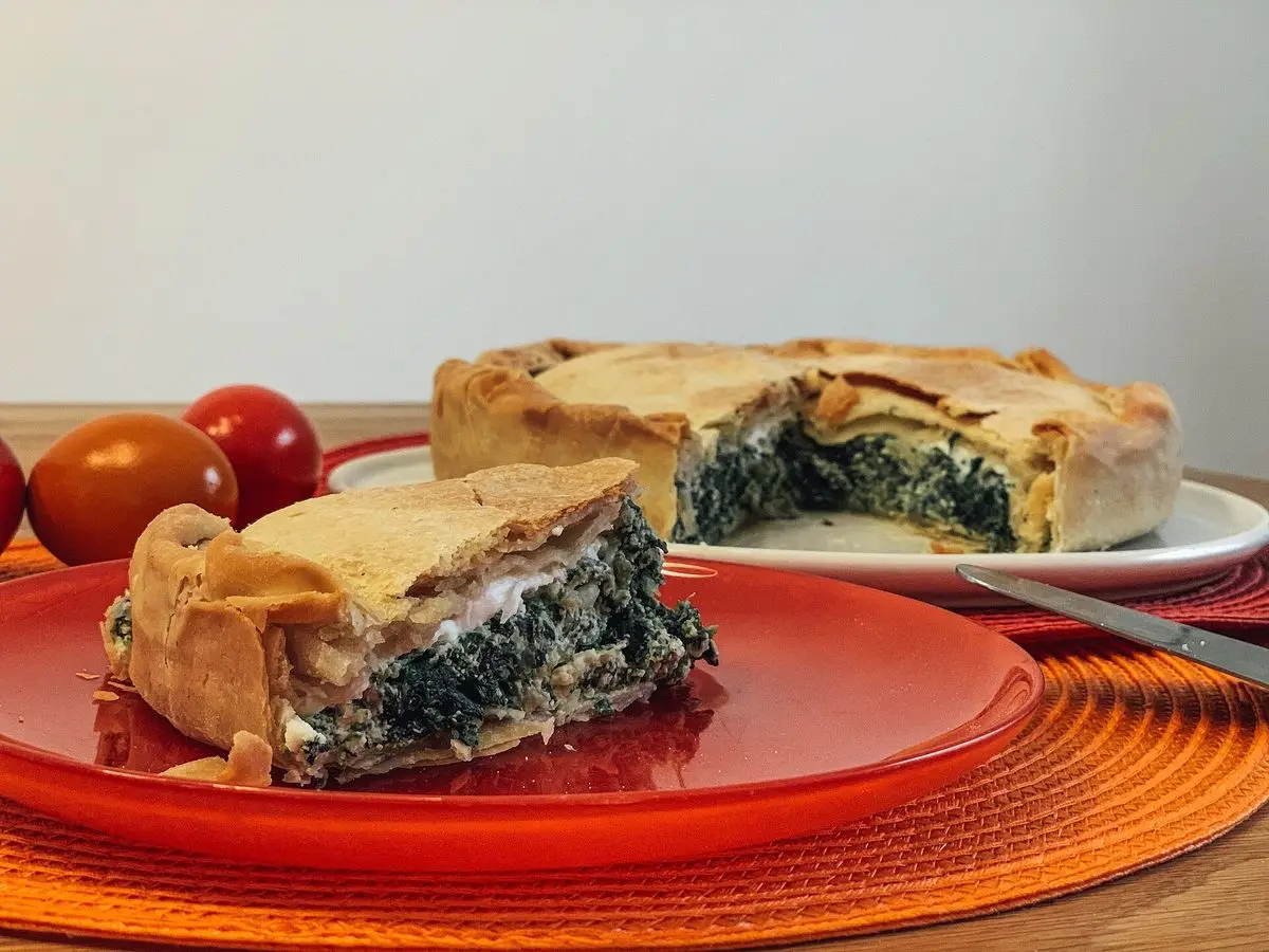 Porción de torta salada de hojaldre fino rellena de espinacas y ricotta en un plato rojo, con la tarta entera detrás