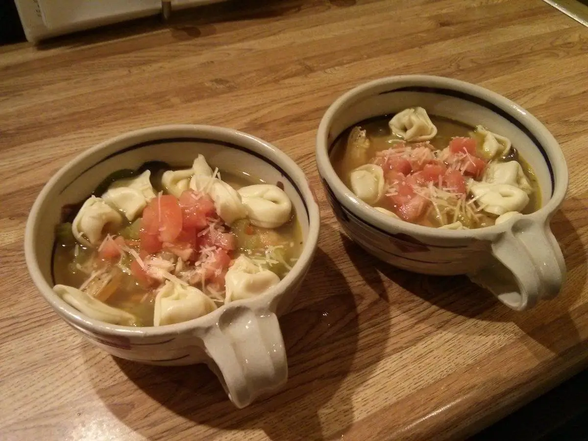 Dos cuencos de sopa de tortellini con pollo y verduras, coronados de tomate picado y queso rallado