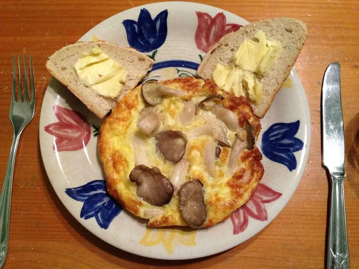 Tortilla abierta de setas laminadas en un plato de flores azules con rebanadas de pan y un tenedor