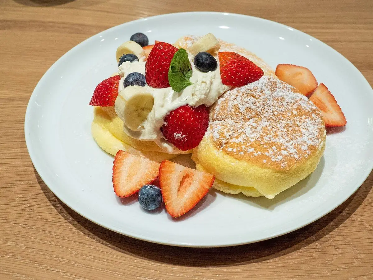 Plato blanco con tortitas japonesas esponjosas, fresas, arándanos, plátano y nata sobre una mesa de madera