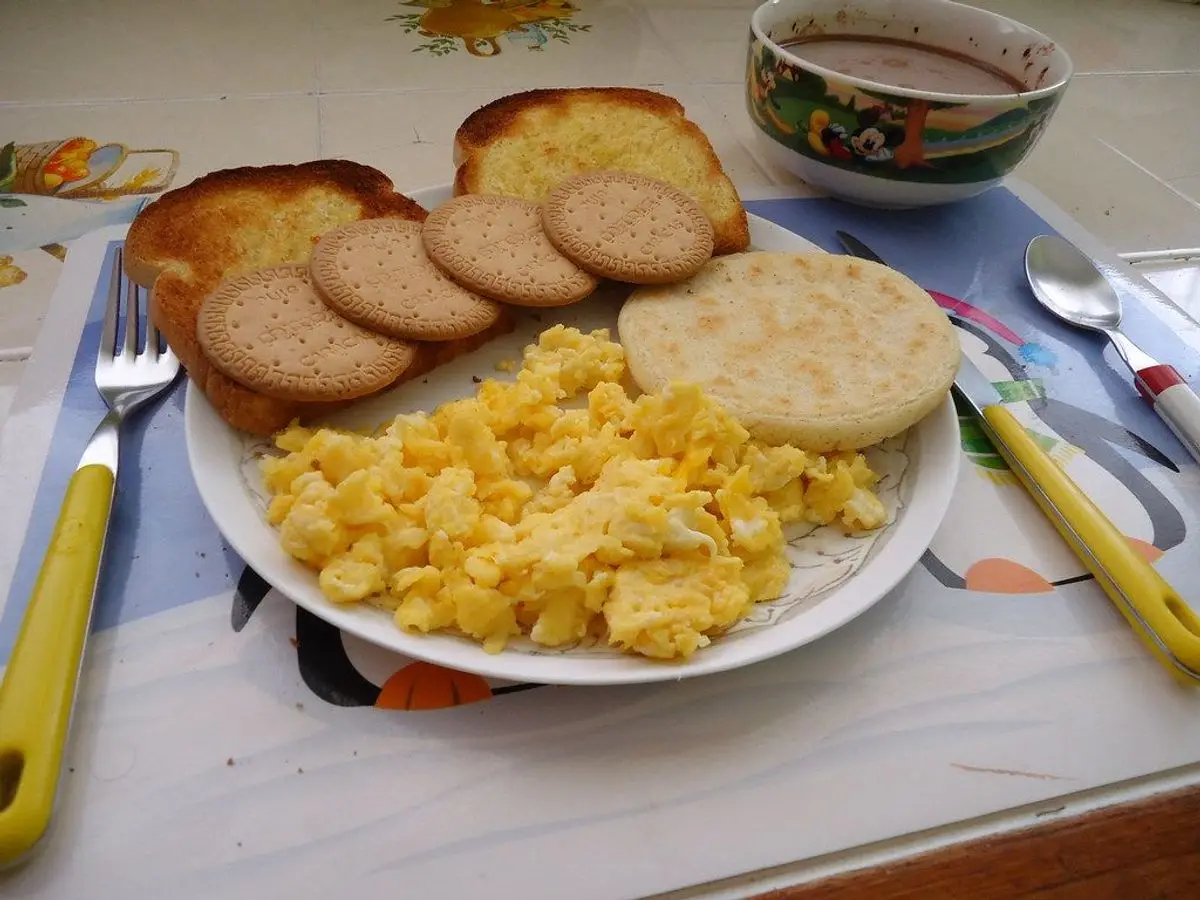 Plato con arepa de maíz, huevo revuelto y rodajas de embutido, con una taza al fondo