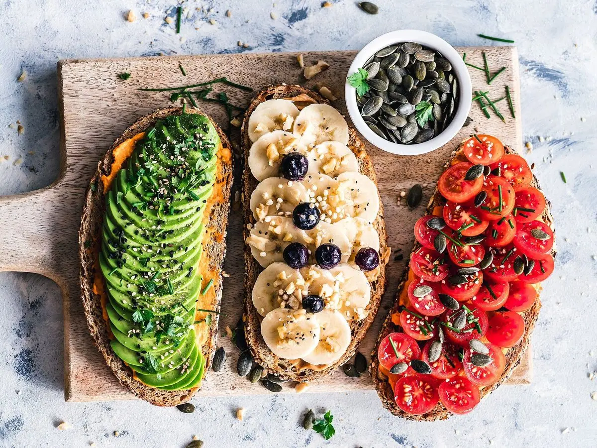 Tres tostas sobre una tabla: aguacate laminado, plátano con arándanos y tomate cherry, con un cuenco de semillas