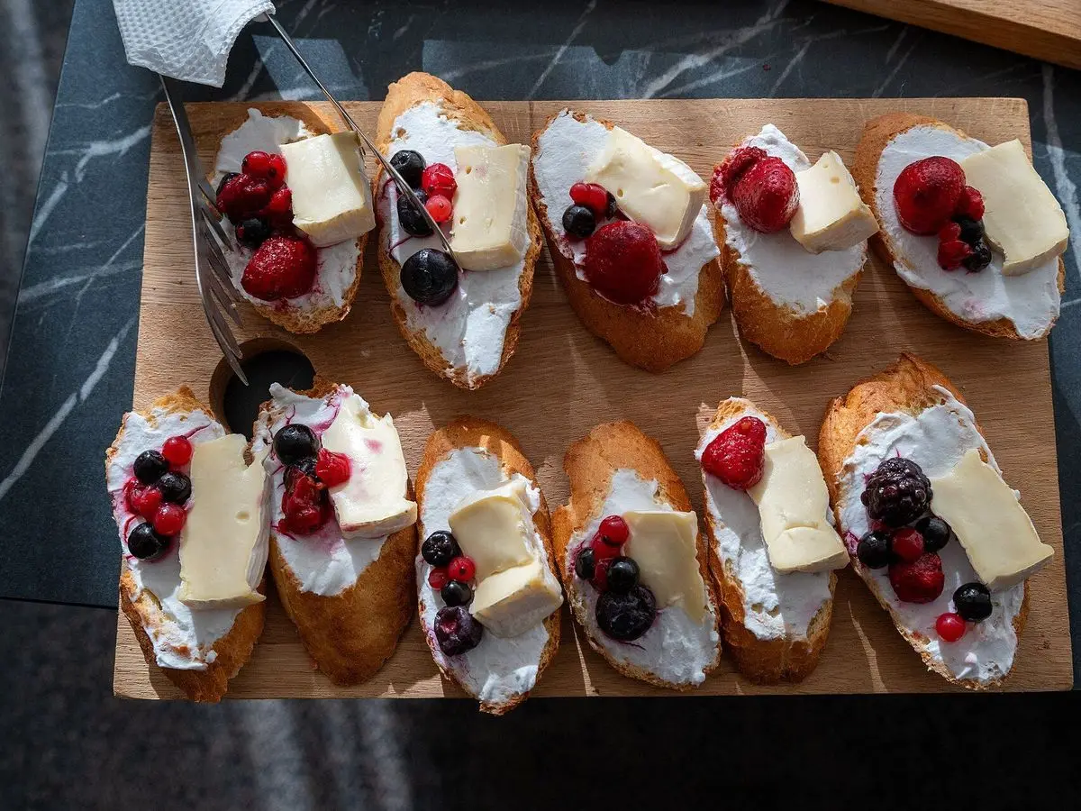 Tostas de pan con queso cremoso blanco, frambuesas, arándanos y láminas de queso sobre una tabla de madera
