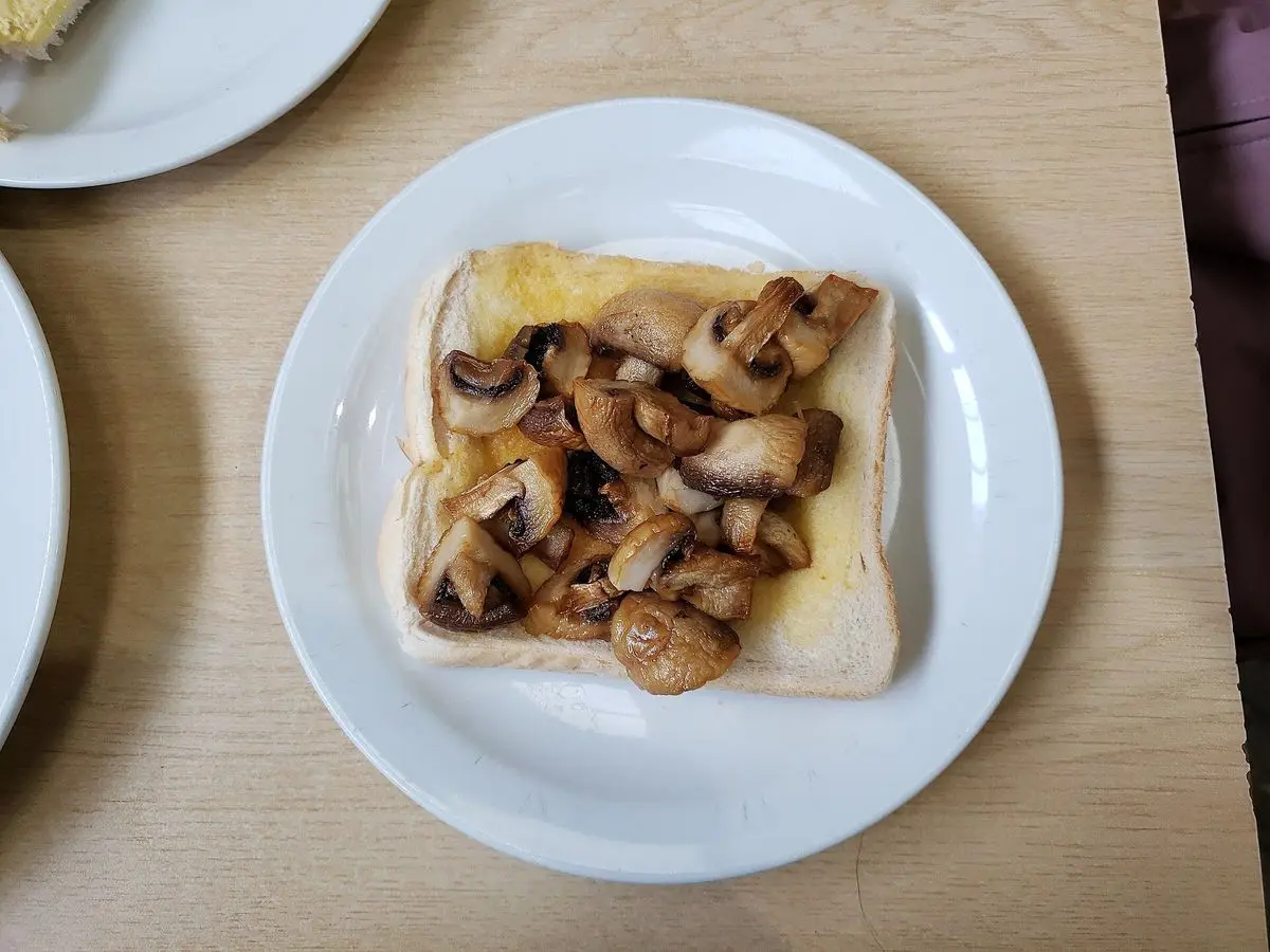 Tostada de pan con champiñones salteados sobre un plato blanco en una mesa de cafetería