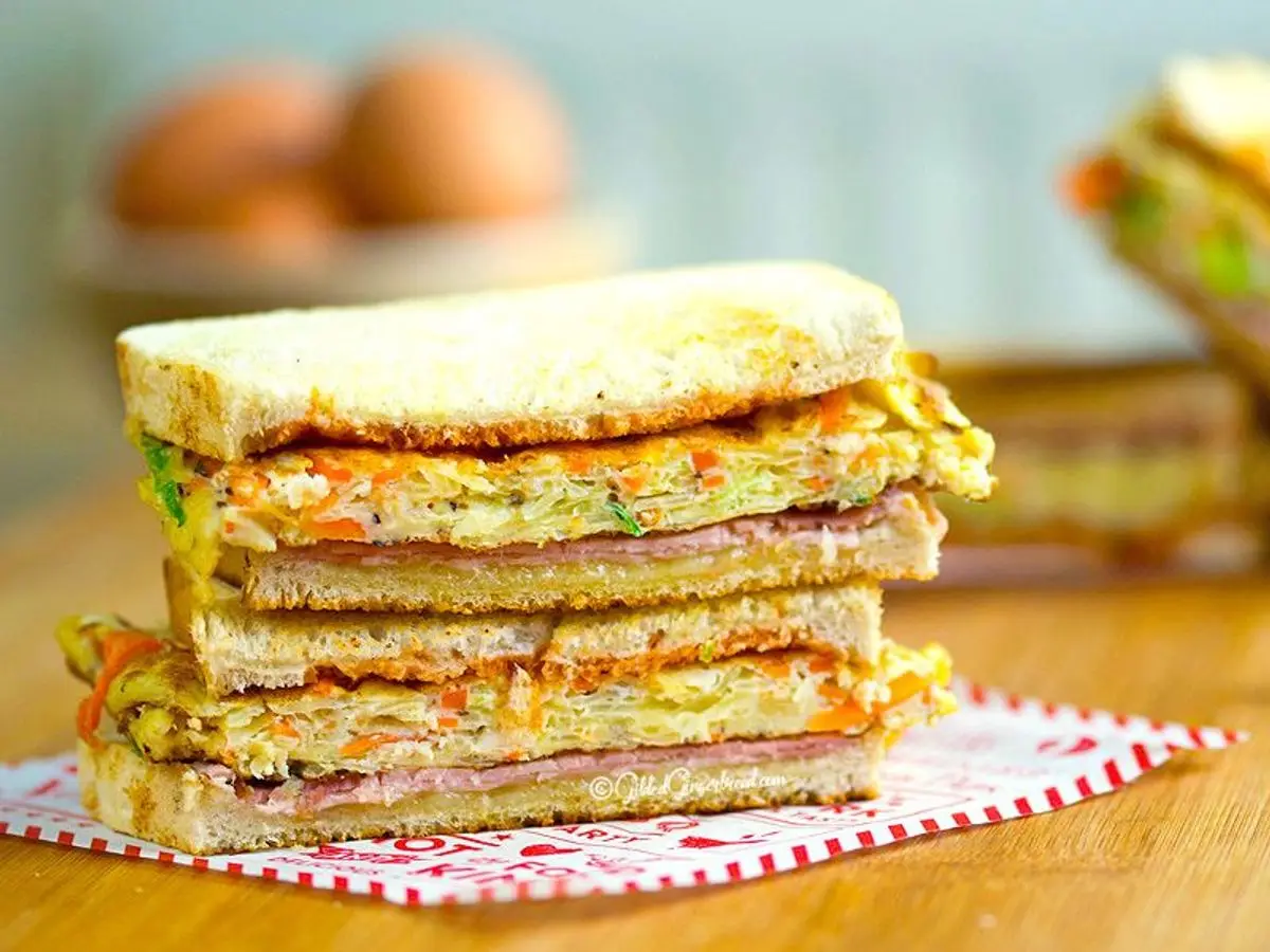 Sándwich de pan tostado con tortilla de verduras y jamón apilado sobre una mesa de madera