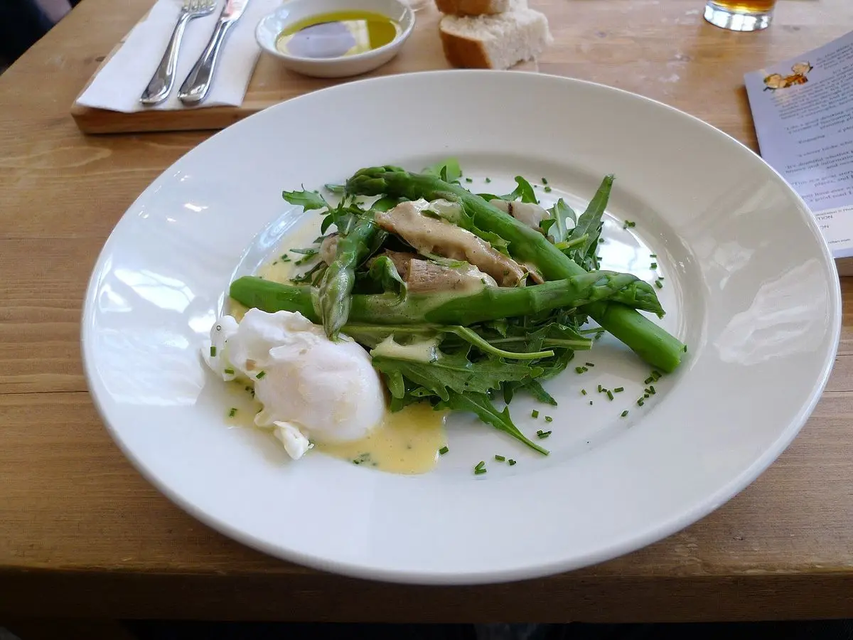 Plato blanco con espárragos verdes, huevo poché y salsa, sobre una mesa de madera