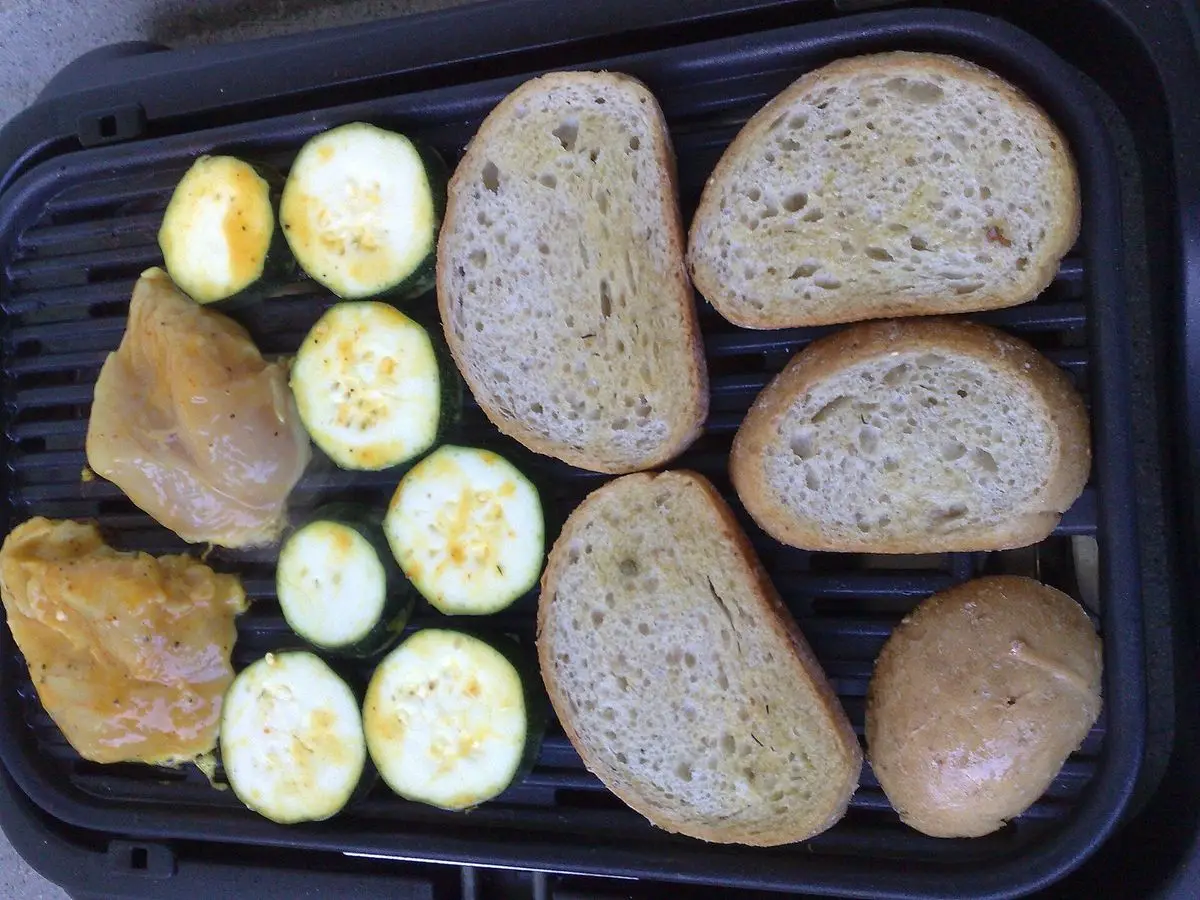 Rodajas de pan y calabacín asándose en una plancha junto a trozos de pollo y patata