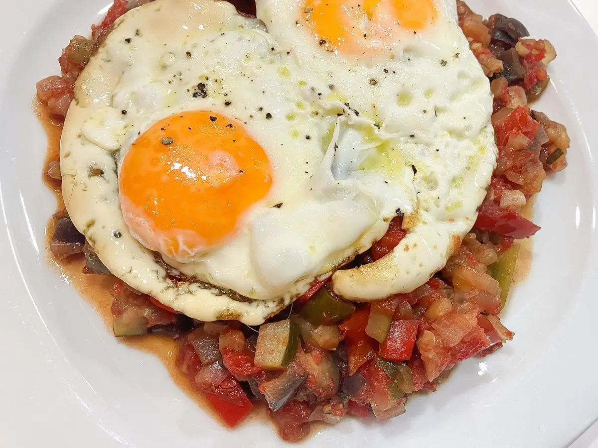 Dos huevos fritos sobre una cama de pisto de verduras en un plato blanco