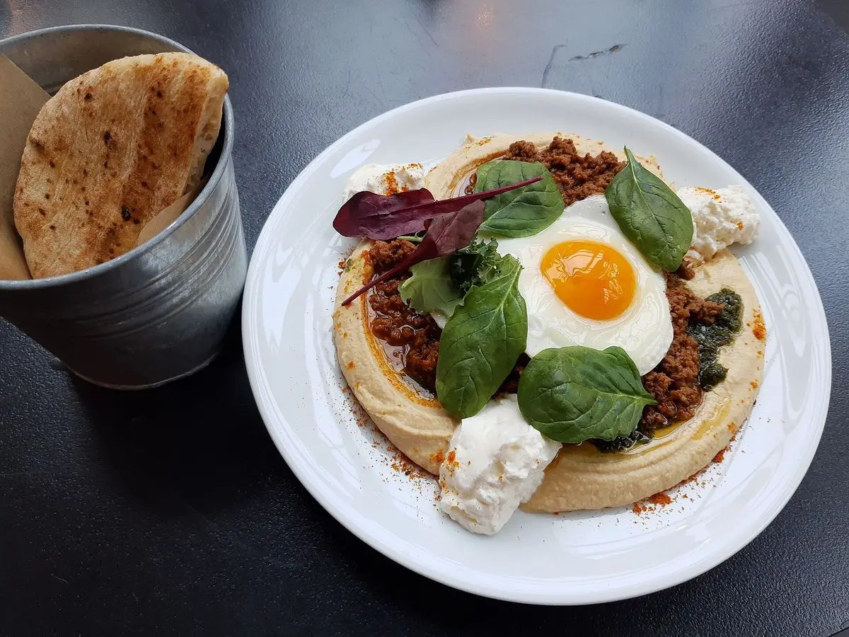 Plato de hummus cremoso con huevo frito, hojas de albahaca y pan de pita al lado