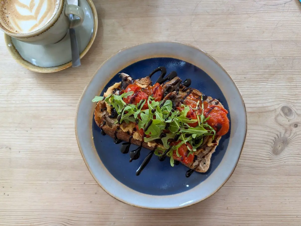 Tostada abierta con hojas verdes, tomate y aliño oscuro en un plato azul junto a un café con leche