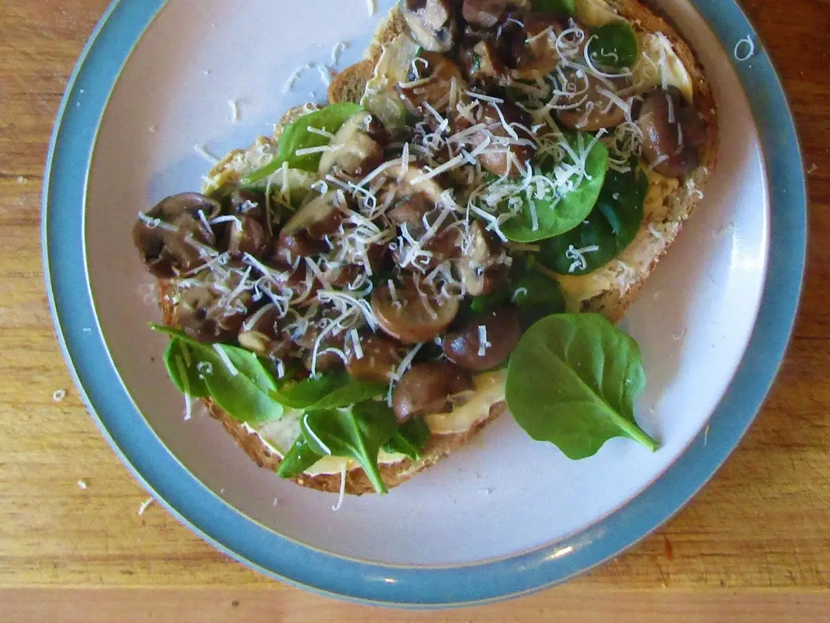 Tostada de pan integral con champiñones, espinacas y queso rallado en un plato con borde azul