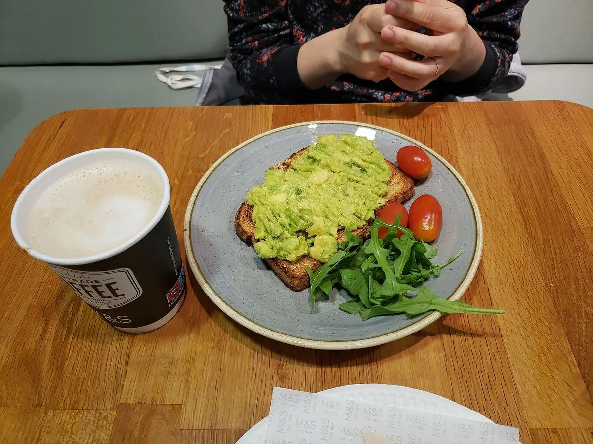 Tostada con aguacate machacado y tomates cherry en un plato con rúcula junto a un café