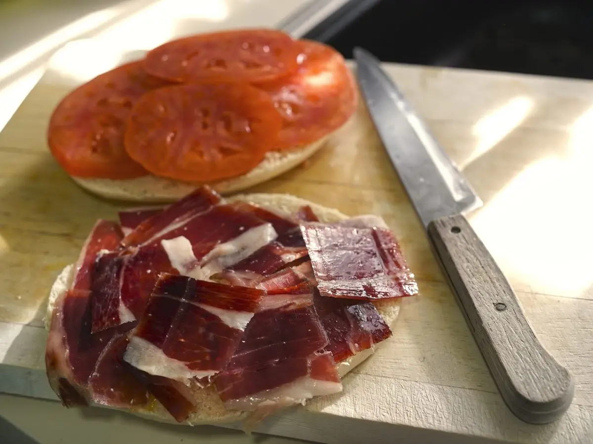 Pan abierto sobre una tabla: una mitad con rodajas de tomate y la otra con lonchas de jamón ibérico