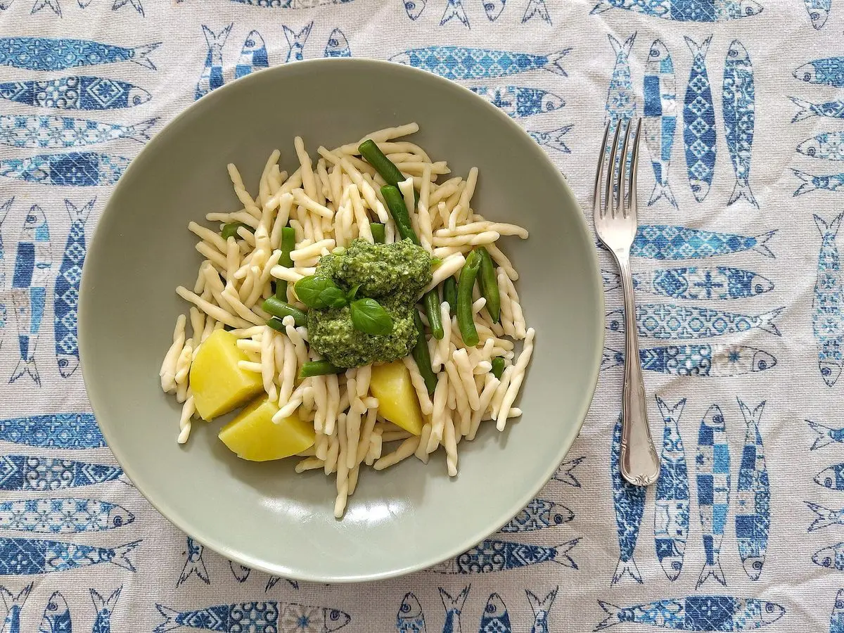 Plato verde de pasta trofie con pesto, trozos de patata y judías verdes sobre un mantel de peces azules