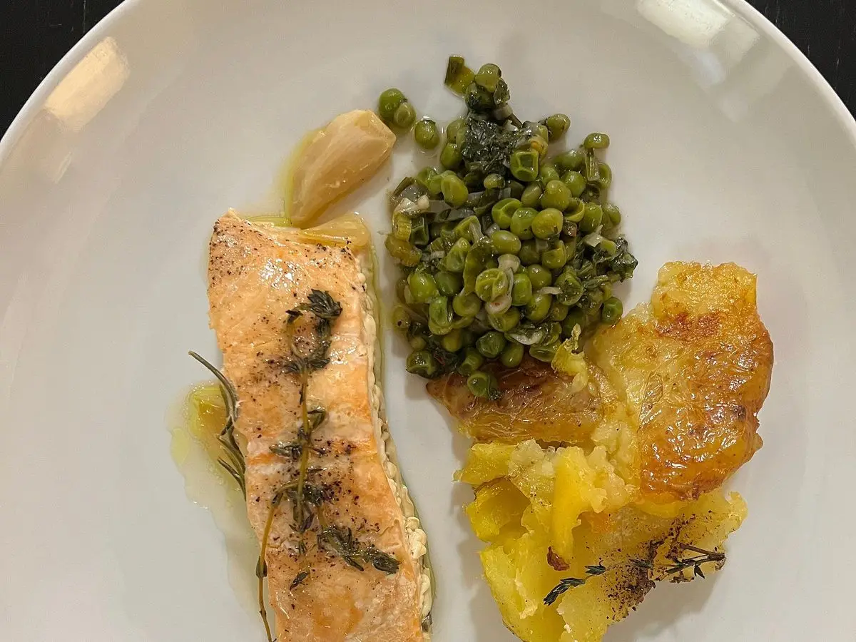 Filete de pescado rosado con hierbas junto a guisantes y patatas doradas en un plato blanco