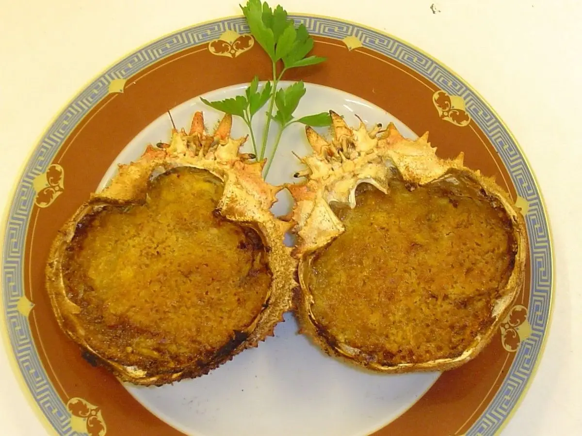 Dos caparazones de centollo rellenos y gratinados dorados sobre un plato con ribete naranja y perejil